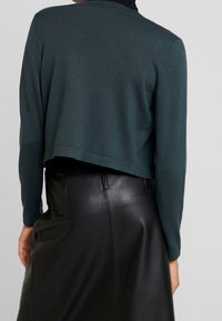 Person wearing a dark green cropped sweater and high-waisted black leather skirt, shown from the back against a plain background.