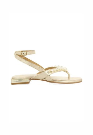 Sandal plano beige con una correa en el tobillo, diseño de tiras cruzadas y acentos de perla en la parte frontal. Detalle metálico en el tacón. Textura suave.