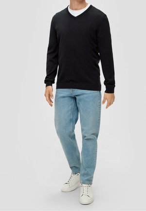 Homme portant un pull à col en V noir sur une chemise blanche, un jean bleu clair et des baskets blanches, debout devant un fond gris.