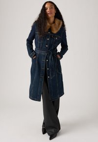 Manteau trench en denim bleu foncé avec un col en fourrure synthétique, taille ceinturée et fermeture à boutons. Il comporte deux poches latérales et une silhouette longue.