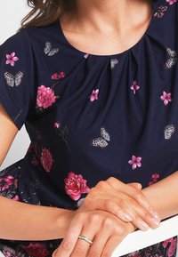 Marinblå topp med korta ärmar, med veckad halsringning och blommönster i form av fjärilar i rosa och grått. Mjuk tygtextur.