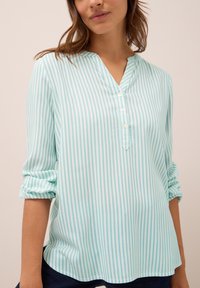 Mujer vestida con una blusa de manga larga a rayas verticales en azul claro y blanco, con tres botones y las mangas arremangadas, combinada con pantalones oscuros.