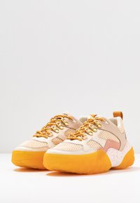 Leichte Sneakers mit beigem Mesh-Obermaterial, goldenen Ösen, orangefarbener Gummisohle und rosa gesprenkelten Akzenten an der Ferse.
