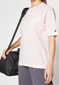 Světle růžové tričko s krátkým rukávem a kulatým výstřihem, se malým logem na hrudi a akcentem loga na rukávu.