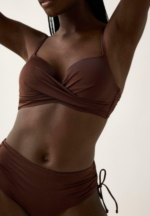 Kvinde iført en mørkebrun todelt badedragt med en omslags-style top og højtaljede underdele med bindebånd i siderne mod en ensfarvet baggrund.