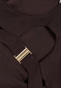 Tumepruun kangas sujuva tekstuuriga, millel on ümmargune ava ja kuldtooni metalldetail, millel on vertikaalsed jooned.