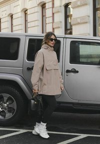 Žena v béžovém kabátě, černých legínách, bílých teniskách a slunečních brýlích, stojící vedle stříbrného zaparkovaného SUV na městské ulici.