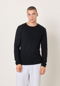 Schwarzer Strickpullover mit rundem Ausschnitt, langen Ärmeln und weicher Textur, kombiniert mit hellgrauen Jogginghosen vor neutralem Hintergrund.