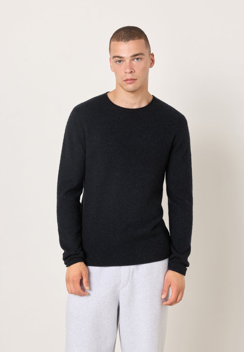 Schwarzer Strickpullover mit rundem Ausschnitt, langen Ärmeln und weicher Textur, kombiniert mit hellgrauen Jogginghosen vor neutralem Hintergrund.