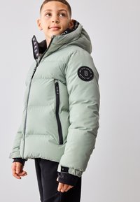 Jachetă de puf verde deschis cu glugă, buzunare cu fermoar, accente negre și un patch cu logo circular pe mânecă. Material texturat.