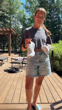 Zapatillas deportivas blancas con detalles verdes, sujetas en las manos de una persona; lleva una camiseta corta gris y pantalones cortos de mezclilla. Entorno en una terraza de madera al aire libre.
