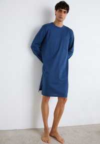 Blå mønstret lang skjorte med rund hals og lange ermer, med brystlomme og splitt foran for ekstra bevegelse. Stoffet ser mykt ut.