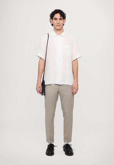 Camisa de lino blanca de manga corta con un bolsillo en el pecho, combinada con pantalones beige ajustados y zapatos negros, sosteniendo un bolso negro pequeño.