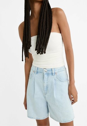 Femme aux longues tresses portant un top tube blanc et un short en jean bleu clair taille haute sur fond blanc.