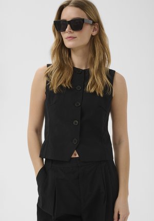 Vrouw met schouderlang blond haar, gekleed in een zwart vestje met knopen, bijpassende broek en grote zwarte zonnebril.