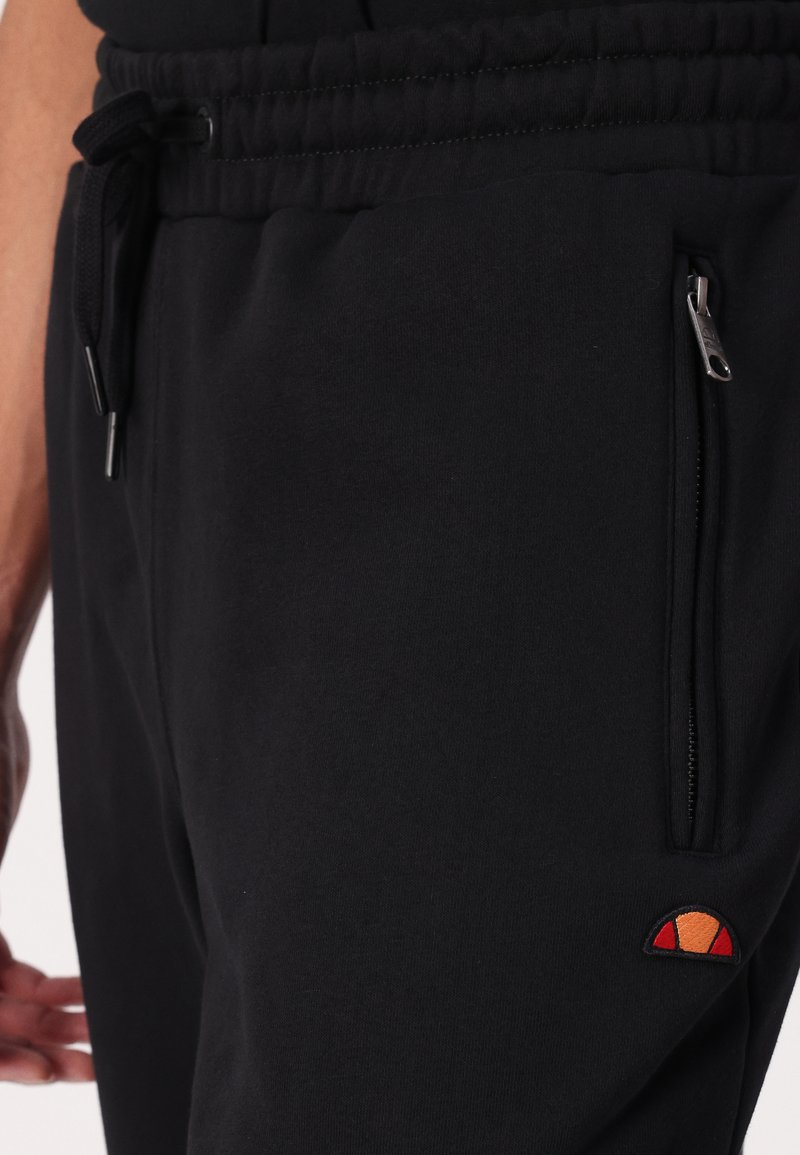 Czarne dresy z wiązaniem w talii, zamykanymi kieszeniami oraz małym haftowanym logo w pomarańczowych i czerwonych akcentach. Gładka tekstura.
