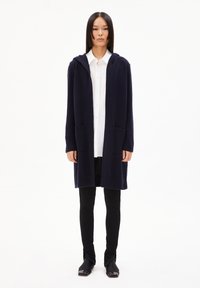 Long gilet en tricot bleu marine avec capuche, deux poches avant et poignets côtelés. Porté par-dessus une chemise blanche à boutons et un legging noir.