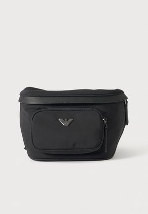 Sac banane noir avec une fermeture éclair sur le dessus, une poche avant zippée et un logo d’aigle argenté, sur un fond clair uni.