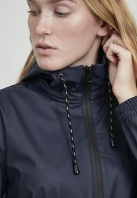 Veste imperméable navy avec un col haut, des cordons de serrage réglables et une fermeture éclair. Texture lisse et tissu durable.