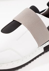 Vit sneaker med ett grått elastiskt band, svarta detaljer, slät yta och en gummisula med små perforeringar och ett zigzag-mönster.