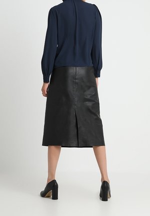 Kvinde iført en marineblå bluse med lange ærmer, sort læder midi-nederdel med slidse bagpå og sorte sko med blokhæl, stående med ryggen til.