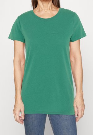 Kvinde iført en ensfarvet grøn kortærmet T-shirt og blå jeans, vist fra nakke til hofter mod en hvid baggrund.