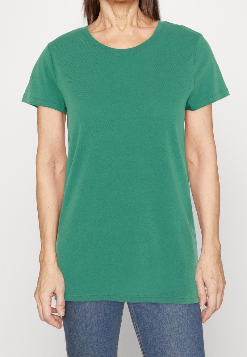 Femme portant un t-shirt vert uni à manches courtes et un jean bleu, vue du cou aux hanches sur fond blanc.
