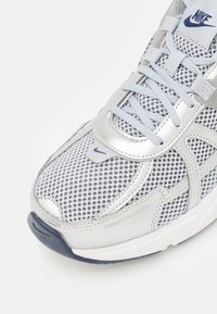 Nike sneaker with a silver and gray mesh upper, textured panels, reflective accents, and a dark blue sole. Features lace closure and logo.