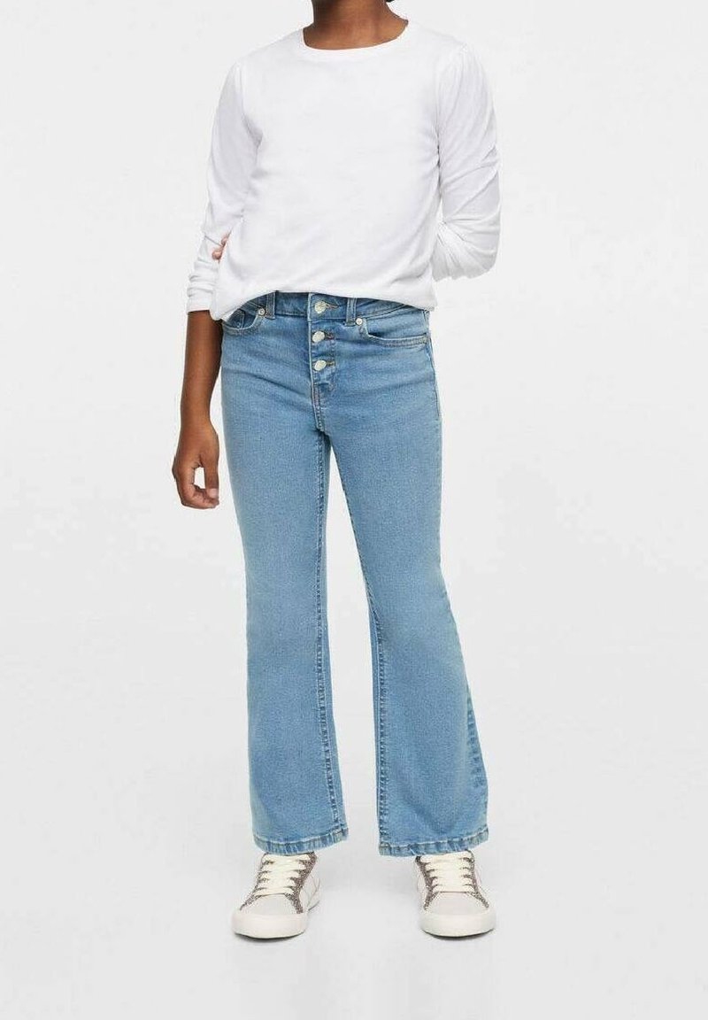 Enfant portant une chemise blanche à manches longues, un jean évasé taille haute bleu clair avec des boutons, et des baskets blanches, debout devant un fond uni.