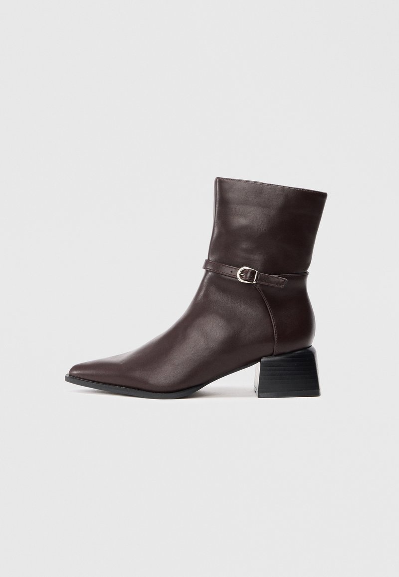 Bottine en cuir marron foncé avec bout pointu, petit talon carré et fine bride avec boucle argentée autour de la cheville.