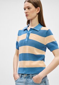 Νεαρή γυναίκα με μακριά μαλλιά που φοράει ένα πλεκτό πόλο μπλουζάκι με κοντά μανίκια σε μπλε και μπεζ ρίγες, με τα χέρια στις τσέπες ανοιχτόχρωμων μπλέ τζιν.