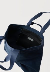 Tumesinine kandekott kahe pika käepidemega ja must lukuga sisetasku, millel on silt Lacoste.