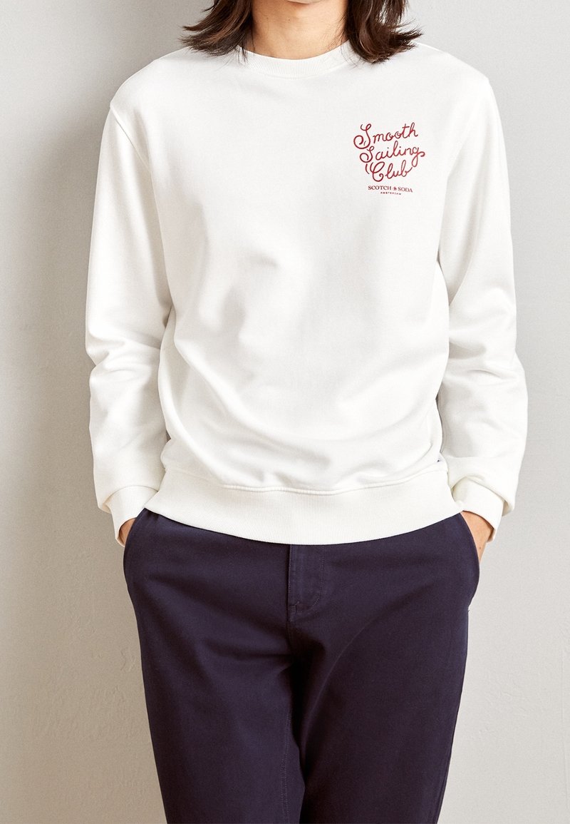 Personne portant un sweat-shirt blanc avec le texte rouge « Smooth Sailing Club » sur la poitrine et un pantalon bleu marine foncé, mains dans les poches, sur fond uni.