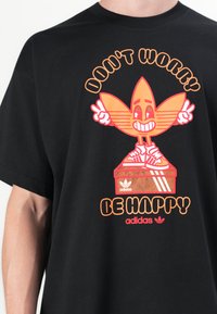 Černé bavlněné tričko s velkým oranžovým kresleným grafikou a textem "NESTAREJ SE, BUĎ ŠŤASTNÝ" a logem Adidas. Široké krátké rukávy.