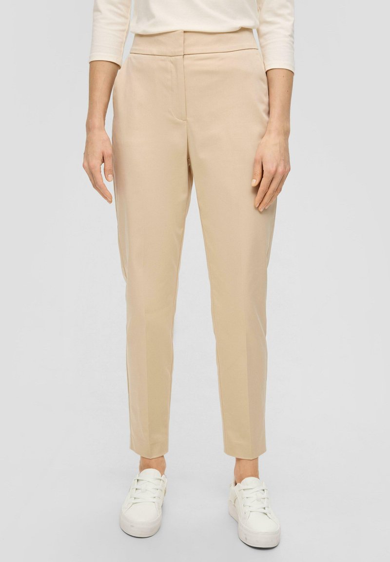 Pantalons beiges ajustés avec une texture lisse, coupe slim et longueur cheville, associés à des baskets blanches. Lignes épurées et design minimal.