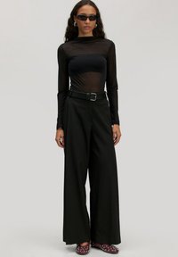 Zwarte doorzichtige lange mouwtop, getailleerd bij de torso. Brede zwarte broek met een riem. Bordeau slip-on schoenen met zilveren studs. Bijpassende donkere zonnebril.