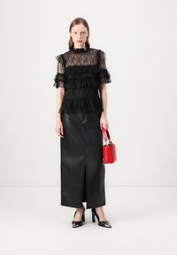 Top in pizzo nero con strati arricciati, abbinato a una lunga gonna in pelle nera con uno spacco frontale. Accessoriato con una piccola borsa rossa.