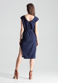 Femme aux longs cheveux bruns portant une robe sans manches bleu marine avec une fente latérale et des sandales à talons marron, debout devant un fond uni.