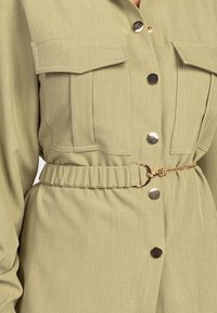 Olive green shirt dress featuring a button-up front, chest pockets, gathered waist with a gold chain belt, and smooth, textured fabric.