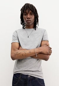 Tricou gri cu mânecă scurtă dintr-un material moale, cu decolteu rotund. Purtați-l cu blugi denim închis și bijuterii din argint, având tatuaje vizibile.