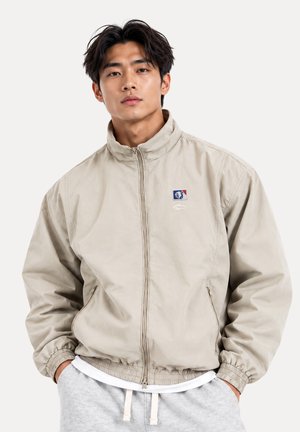 Jeune homme aux cheveux foncés portant une veste beige zippée et un pantalon de survêtement gris clair, les mains dans les poches, sur un fond clair.
