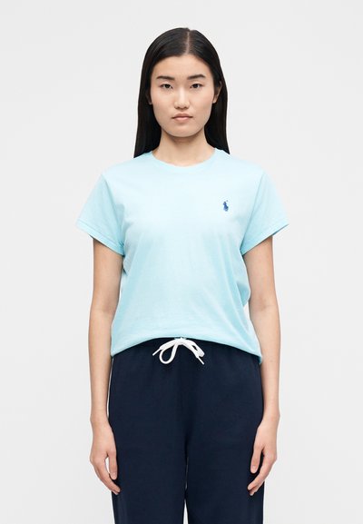 Sieviete ar gariem tumšiem matiem, ģērbusies gaiši zilā īsroku t-kreklā un tumši zilos biksēs ar auklu, stāv pret vienkāršu gaišu fonu.