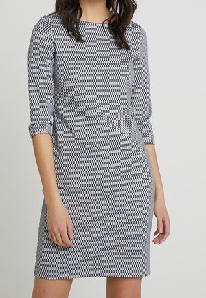 Mujer con un vestido de manga larga hasta la rodilla, con un patrón geométrico en blanco y negro, sobre un fondo gris liso.