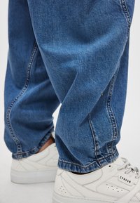 Μπλε τζιν από τζιν ύφασμα με χαλαρή εφαρμογή και ελαστικές μανσέτες, συνδυασμένο με λευκά αθλητικά παπούτσια με διατρητικές λεπτομέρειες και λογότυπο.