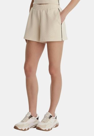 Pantaloncini beige con fascia elastica in vita, rifiniture nere e tasche laterali, indossati con scarpe sportive bianche e beige da una persona in piedi.