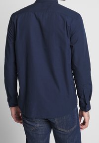 Maglietta a maniche lunghe di colore blu navy con tessuto testurizzato, polsini con bottoni e colletto classico. Vista posteriore che mostra un orlo dritto e dettagli sottili.