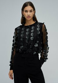 Blouse noire avec des manches transparentes, ornée d'appliqués en forme de fleurs en tissu. Un pantalon noir complète la tenue. Encolure ronde simple.