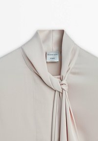 Blouse rose clair avec un nœud au col, fabriquée dans un tissu lisse. L'étiquette indique "Massimo Dutti Studio." Lignes épurées et texture douce.