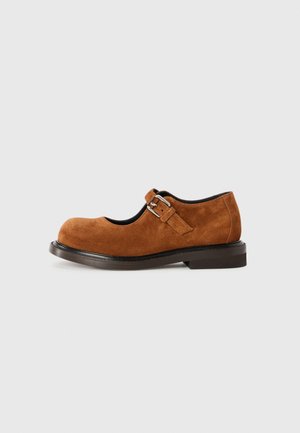 Zapato Mary Jane de ante marrón con punta redonda, presenta un detalle de hebilla lateral y suela de goma plana. Textura suave y líneas limpias.