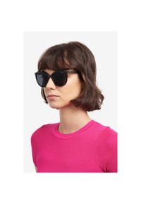 Occhiali da sole neri a forma di occhio di gatto con lenti scure e un sottile accento dorato sulle aste, tenuti da una persona che indossa una camicia rosa aderente.